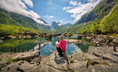 Person i rosa genser ved siden av ein hund. Dei sit ved ein kai, ved irrgrønt brevatn. Grønkledde fjellsider og blå himmel. Foto.