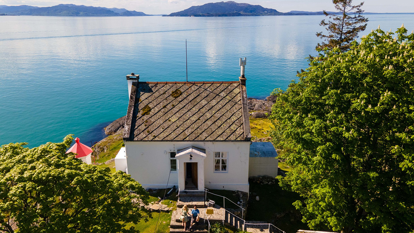 Kvitt murhus med fyrlykt ved sida av seg. Leirvik Fyr. Irrgrøn sjø, grøne lauvtre og nokre øyer i horisonten. Foto.