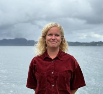 Foto av person i raud skjorte, og svart skjørt som står på platting ved sjø og fjell.