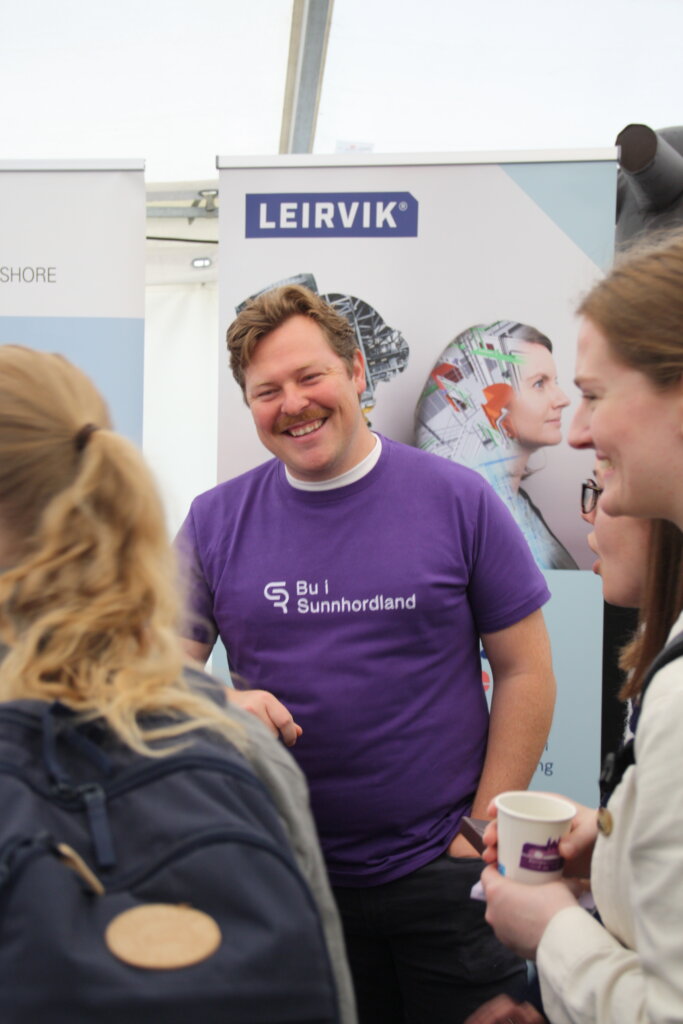 Person iført lilla tskjorte med tekst Bu i Sunnhordland smiler og snakkar med 3 andre personar som også smiler