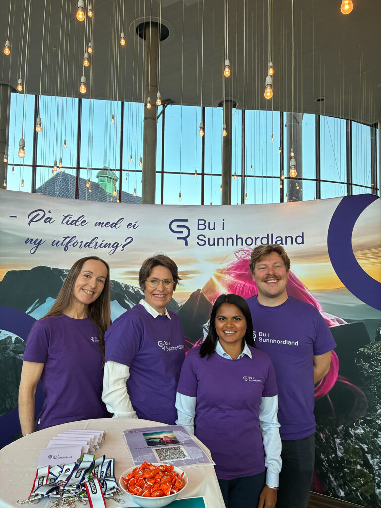 Fire personar iført lilla t-skjorter med tekst Bu i Sunnhordland foran messevegg.