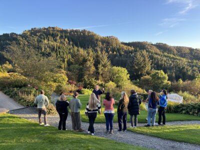 Ei gruppe menneske står samla i ein vakker hage omgitt av haustfarga tre og grøne åsar, medan ein guide formidlar noko. Klår hausthimmel og sollys set stemninga i det frodige landskapet.