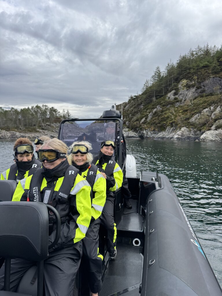 Ei gruppe personar i gule og svarte flytedrakter og vernebriller sit i ein RIB-båt på sjøen, omgitt av holmar og skogkledde berg under overskya himmel.