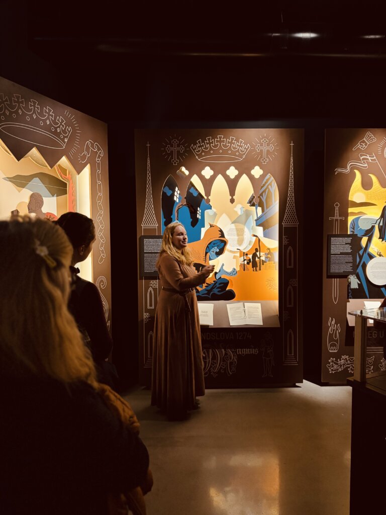 Ei kvinne i historisk drakt formidlar foran ei fargerik og illustrert utstillingsvegg med middelaldermotiv, medan ei gruppe lyttar. Biletet er teke innandørs med dempa belysning.