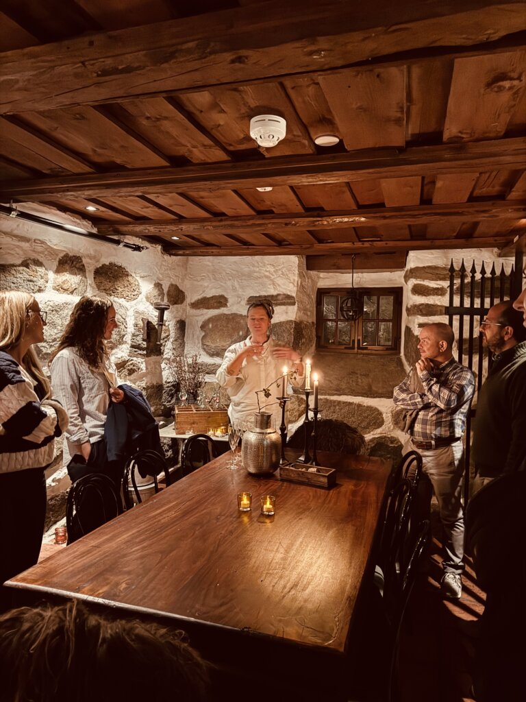 Ei gruppe menneske står samla rundt eit langbord i eit rom med tjukke steinveggar og lågt tretak. På bordet står levande lys og ei stor mugge, medan ein guide fortel engasjert.