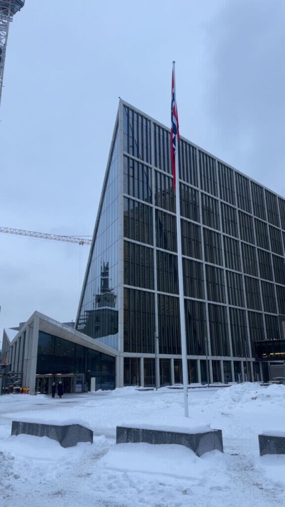 Foto av del av det nye regjeringskvartalet, flaggstang med norsk flagg i, snødekt utenfor.